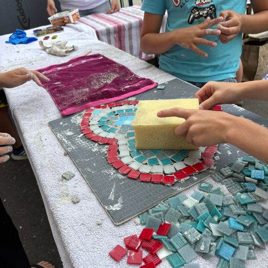 Mehrere Personen arbeiten an einem Mosaikprojekt. Ein Kind hält einen Schwamm und wendet ihn auf ein Mosaik auf einem Tisch an. Das Mosaik hat rote, blaue und grüne Fliesen.