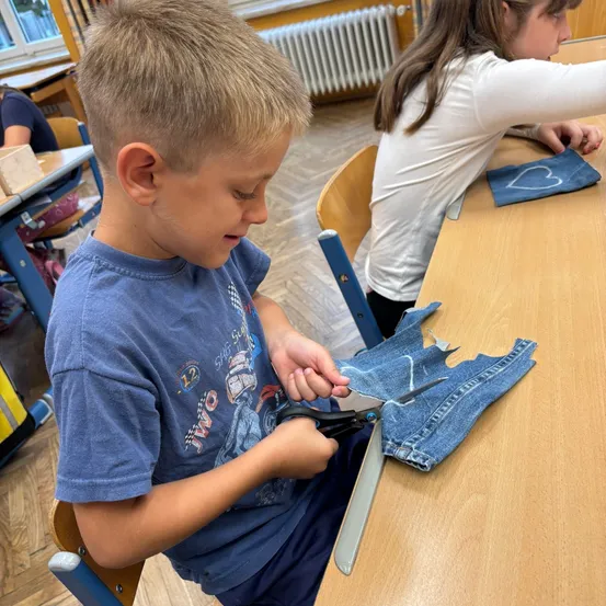 Ein junger Junge sitzt an einem Schreibtisch und schneidet ein Stück Denim-Stoff mit einer Schere. Ein Mädchen in einem weißen Shirt sitzt neben ihm. Hinter ihnen sind weitere Schreibtische und eine Heizung.
