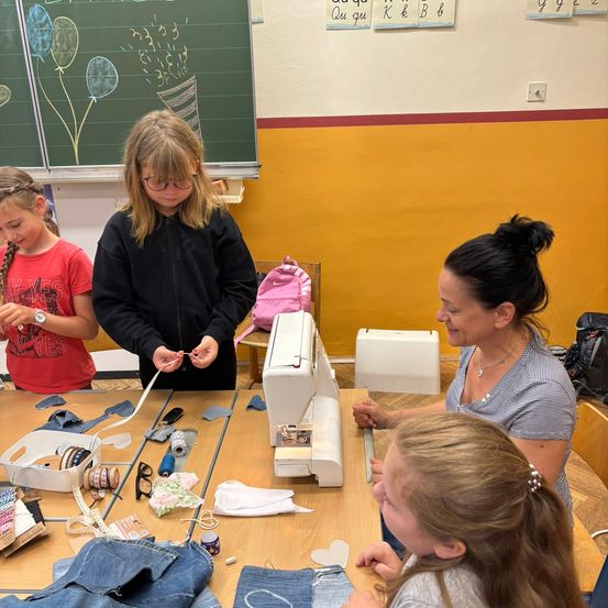 Drei junge Mädchen und eine Frau befinden sich in einem Klassenzimmer. Die Frau unterrichtet die Mädchen im Umgang mit einer Nähmaschine. Der Tisch ist mit Stoff, Scheren und anderen Nähwerkzeugen gefüllt. Hinter ihnen steht eine Tafel mit Zeichnungen von Luftballons.