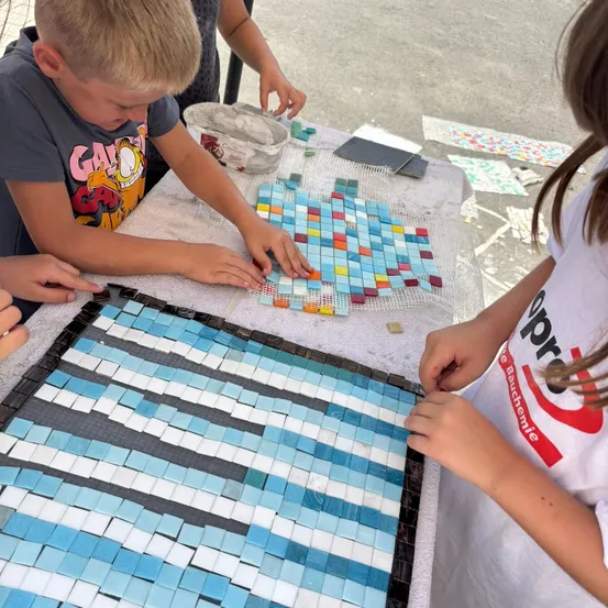 Kinder arbeiten an einem Mosaikprojekt. Sie verwenden verschiedene farbige Fliesen auf einem mit einem weißen Tuch bedeckten Tisch.