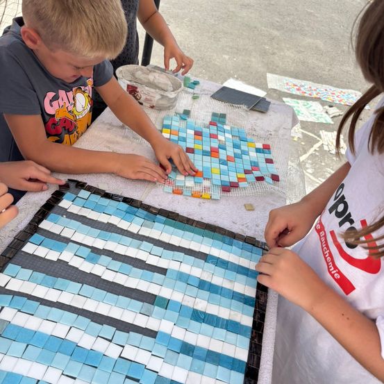 Kinder arbeiten an einem Mosaikprojekt. Sie verwenden verschiedene farbige Fliesen auf einem mit einem weißen Tuch bedeckten Tisch.