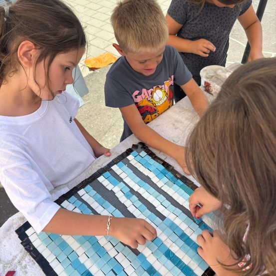 Mehrere Kinder arbeiten an einer Mosaiktafel. Ein Mädchen links trägt ein weißes T-Shirt, während ein Junge in der Mitte ein graues T-Shirt trägt. Ein weiteres Mädchen hinter ihm trägt ein graues T-Shirt mit Punkten.