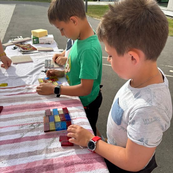 Zwei Jungen arbeiten an einem farbenfrohen Mosaikprojekt auf einem mit einem gestreiften Tuch bedeckten Tisch. Ein Junge trägt ein grünes T-Shirt und eine Uhr, der andere ein graues T-Shirt und eine Halskette.