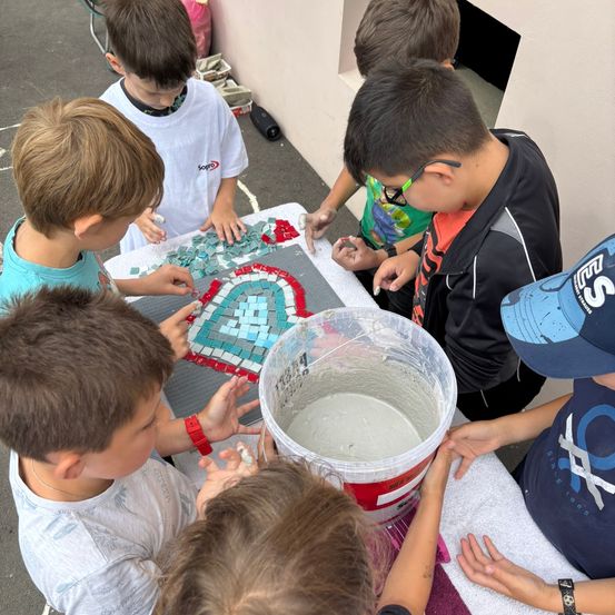 Eine Gruppe von Kindern arbeitet an einem Mosaikprojekt und verwendet farbige Fliesen und Zement. Sie sind um einen Tisch im Freien versammelt, mit einem Eimer Zement in der Mitte.