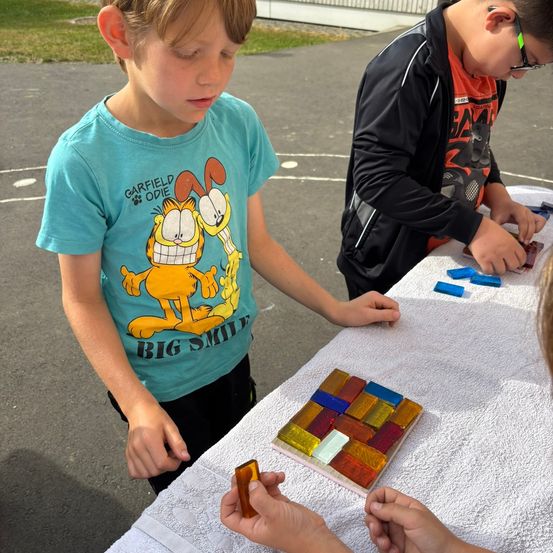 Zwei Kinder arbeiten an einem farbenfrohen Glasmosaik. Ein Junge in einem Garfield-Shirt beobachtet, während der andere in einem schwarzen Sweatshirt Stücke hinzufügt.