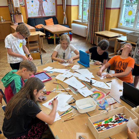 Mehrere Kinder sitzen um einen Tisch herum und zeichnen mit Buntstiften auf Papier. Ein Kind benutzt ein Tablet. Sie sind in einem Klassenzimmer mit Holztischen, Fenstern und einem Bücherregal.