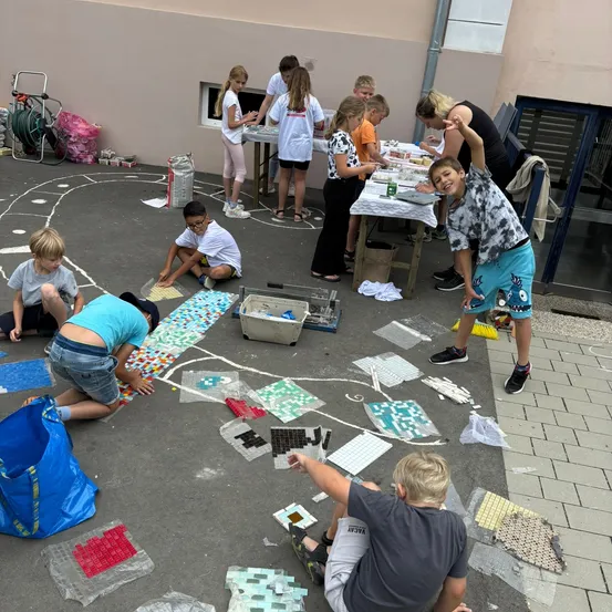 Kinder arbeiten an einem Mosaikprojekt am Boden, tragen Shorts und T-Shirts. In der Nähe sitzen eine Frau und einige Kinder an einem Tisch mit verschiedenen Materialien. Ein Gebäude mit einem Fenster und ein Trolley sind im Hintergrund zu sehen.