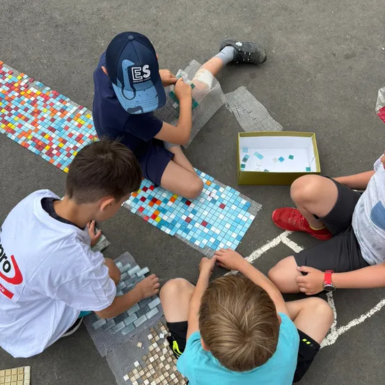 Vier Jungen sitzen auf dem Boden und beschäftigen sich mit einem Bastelprojekt. Einer legt Fliesen auf ein Mosaik, während ein anderer eine Schachtel mit Fliesen hält. Der Junge mit dem blauen Hut schaut auf eine Karte. Der Junge im roten Schuh hält die Hand eines Jungen.