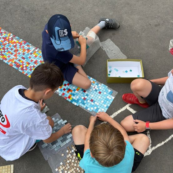 Vier Jungen sitzen auf dem Boden und beschäftigen sich mit einem Bastelprojekt. Einer legt Fliesen auf ein Mosaik, während ein anderer eine Schachtel mit Fliesen hält. Der Junge mit dem blauen Hut schaut auf eine Karte. Der Junge im roten Schuh hält die Hand eines Jungen.
