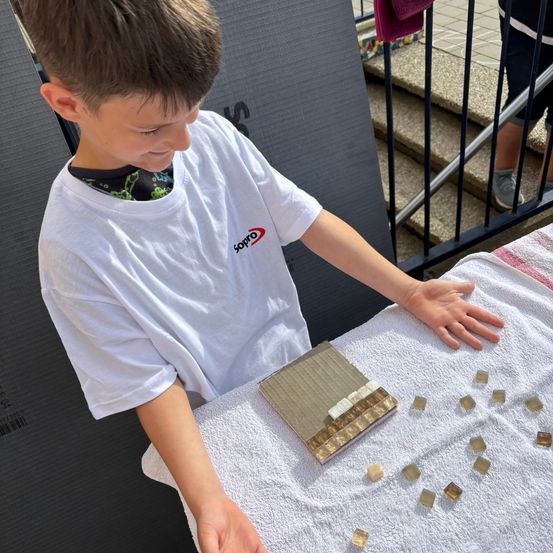 Ein junger Junge in einem weißen T-Shirt ordnet kleine quadratische Fliesen auf einem Handtuch an. Die Fliesen sind um ihn herum auf dem Tisch verstreut. Er schaut auf die Fliesen hinunter.