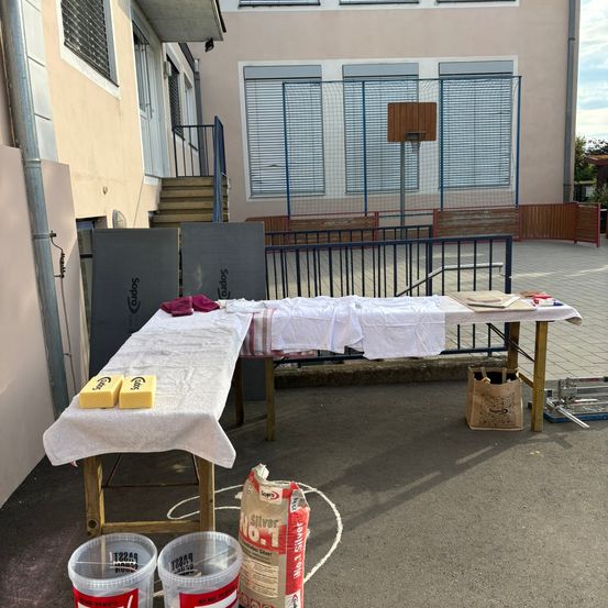 Ein Tisch mit einem weißen Tuch und Reinigungswerkzeugen ist im Freien aufgestellt. Ein Basketballkorb befindet sich hinter dem Tisch. Eimer und ein Sack Zement liegen auf dem Boden.