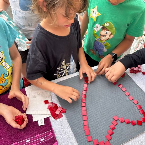 Vier Kinder arbeiten zusammen, um ein herzförmiges Mosaik mit roten Fliesen auf einem grauen Untergrund zu gestalten. Jedes Kind hält Fliesen, und zwei platzieren sie. Ein Kind trägt ein schwarzes T-Shirt mit einem weißen Design.