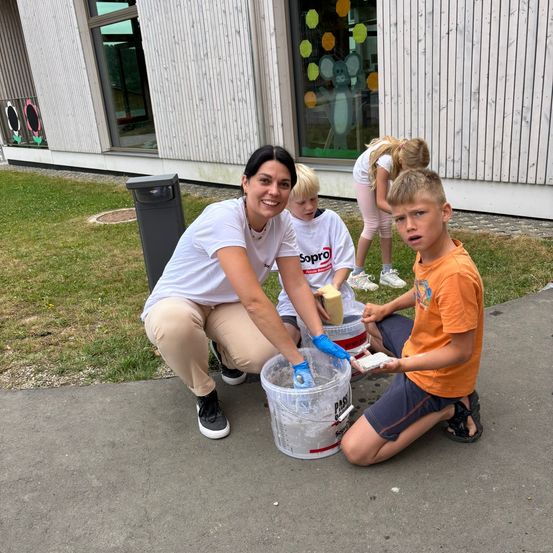 Eine Frau und drei Kinder sind auf einem Bürgersteig mit Farbbechern. Ein Kind hält einen Schwamm und ein anderes einen Pinsel. Ein Gebäude mit Glasfenstern ist im Hintergrund.