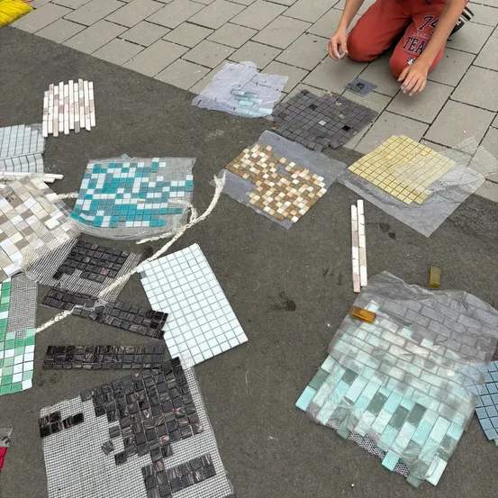 Eine Person kniet auf einem Bürgersteig mit verschiedenen verstreuten Fliesen, um eine Mosaik zu verlegen. Es gibt mehrere Farben und Größen von Fliesen auf dem Boden, und einige sind in Plastik eingewickelt.