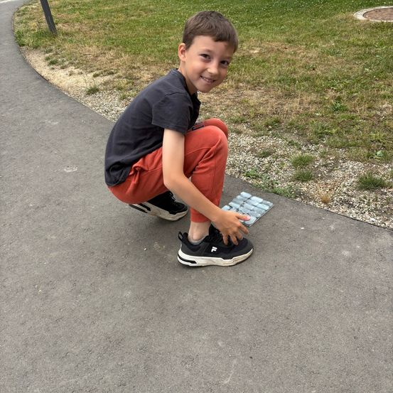 Ein Junge in einem schwarzen Shirt und roten Hosen kniet auf einem gepflasterten Weg und untersucht lächelnd ein kleines quadratisches Mosaik auf dem Boden.