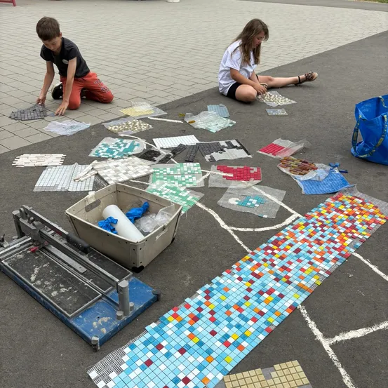 Zwei Kinder sitzen auf dem Boden, umgeben von bunten Fliesen, und scheinen an einem Mosaikprojekt zu arbeiten.