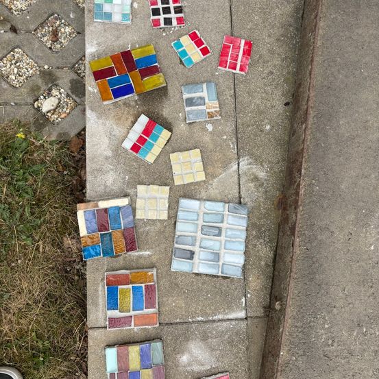 Eine Auswahl an farbenfrohen Mosaikfliesen, die auf einem Betonpflaster neben Gras und Bordstein verstreut sind.