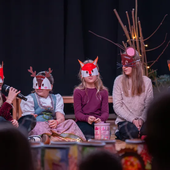 Vier Kinder mit Masken sitzen auf der Bühne, eines hält ein Mikrofon. Sie tragen Kostüme und sitzen zwischen Dosen und Zweigen. Hinter ihnen ist ein schwarzer Hintergrund.