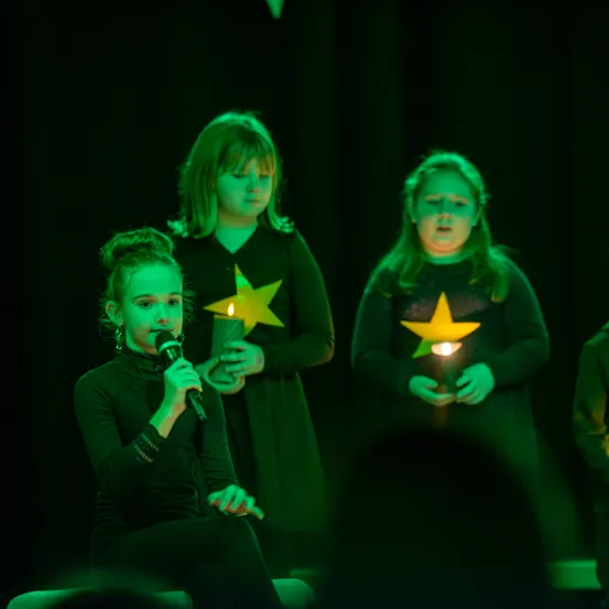 Drei Mädchen treten auf der Bühne auf, eine sitzt und singt in ein Mikrofon, eine andere hält eine brennende Kerze und die dritte steht dahinter mit einer brennenden Kerze. Sie tragen passende Outfits mit gelben Sternen.
