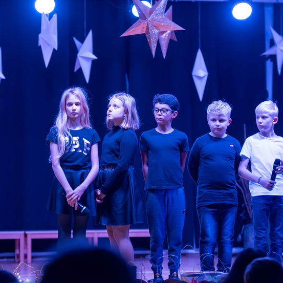 Fünf Kinder stehen auf der Bühne, drei Mädchen und zwei Jungen, mit Mikrofonen, unter einem blauen Hintergrund mit Papiersternen. Die Mädchen tragen schwarze Kleider, während die Jungen dunkle blaue Hemden und Jeans tragen. Der Junge auf der rechten Seite hält ein schwarzes Mikrofon.