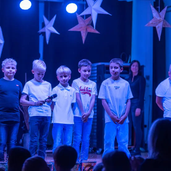 Fünf junge Jungen stehen auf einer Bühne, einer hält ein Mikrofon. Sie sind in weißen T-Shirts und blauen Jeans gekleidet. Hinter ihnen steht eine Frau, und die Bühne ist mit sternförmigen Lichtern dekoriert. Das Publikum sitzt unten und beobachtet die Aufführung.