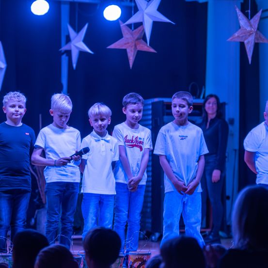 Fünf junge Jungen stehen auf einer Bühne, einer hält ein Mikrofon. Sie sind in weißen T-Shirts und blauen Jeans gekleidet. Hinter ihnen steht eine Frau, und die Bühne ist mit sternförmigen Lichtern dekoriert. Das Publikum sitzt unten und beobachtet die Aufführung.