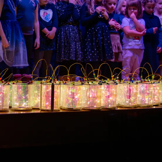 Kinder stehen hinter beleuchteten Glasgläsern auf der Bühne und werfen Licht auf das Publikum. Sie tragen festliche Kleider und scheinen engagiert zu sein.