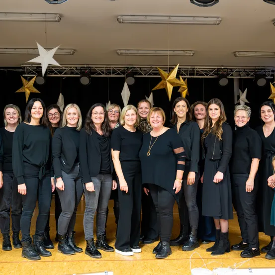 Eine Gruppe von Frauen in schwarzen Kleidern steht in einem Raum mit Holzboden und lächelt für ein Foto. Hinter ihnen hängen goldene Sterndekorationen an der Decke.
