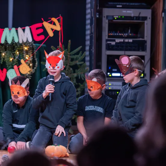 Vier Kinder in Fuchsmasken sitzen auf einer Bühne, eines hält ein Mikrofon. Ein Banner mit bunten Buchstaben hängt hinter ihnen.