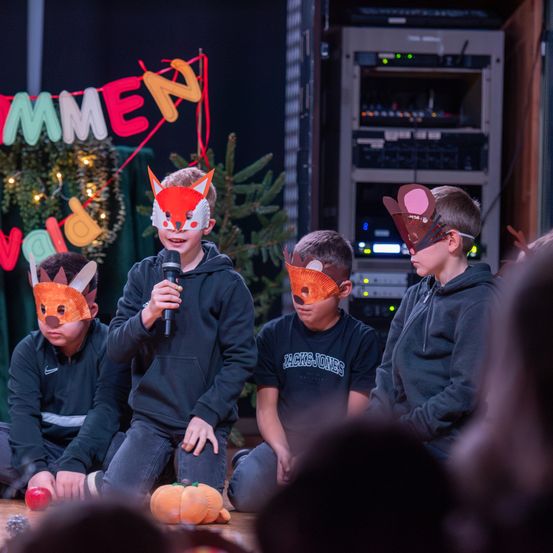 Vier Kinder in Fuchsmasken sitzen auf einer Bühne, eines hält ein Mikrofon. Ein Banner mit bunten Buchstaben hängt hinter ihnen.