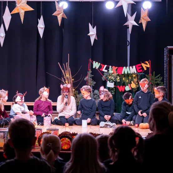 Eine Gruppe von Kindern in Fuchsmasken führt ein Theaterstück auf der Bühne auf. Ein Banner mit der Aufschrift 'Willkommen' hängt über ihnen. Das Publikum beobachtet von vorne.