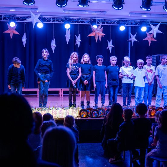 Eine Gruppe von Kindern steht auf einer Bühne mit leuchtenden Laternen vor ihnen und führt vor einem Publikum auf. Sie stehen in einer Reihe, wobei einige Sterne von der Decke hängen.