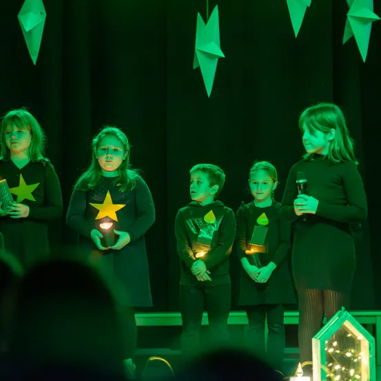 Fünf Kinder stehen auf der Bühne bei einer Aufführung, beleuchtet von grünen Lichtern. Sie tragen sternenförmige Kleidung und halten leuchtende Laternen. Ein Mädchen auf der rechten Seite hält ein Mikrofon.