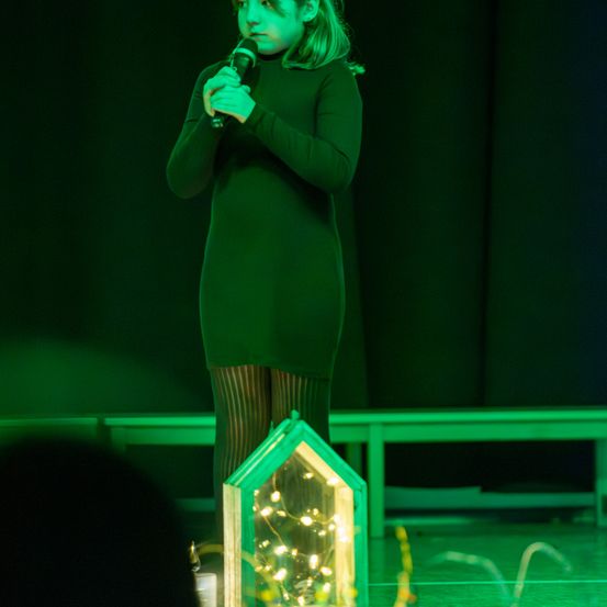 Ein junges Mädchen in einem grünen Kleid steht auf der Bühne, hält ein Mikrofon in der Hand und tritt auf. Vor ihr steht ein dekoratives Haus mit brennenden Kerzen.