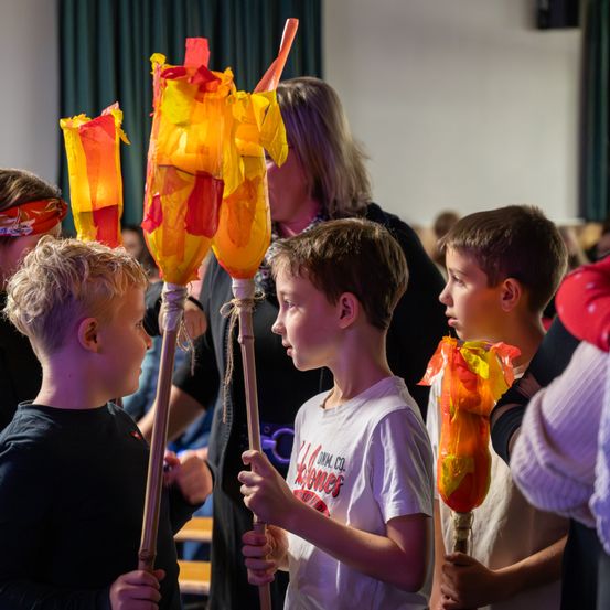 Vier Jungen halten bunte Laternen in einem Raum. Der Junge auf der rechten Seite trägt ein weißes T-Shirt mit rotem Text. Eine Frau mit blondem Haar steht hinter ihnen.