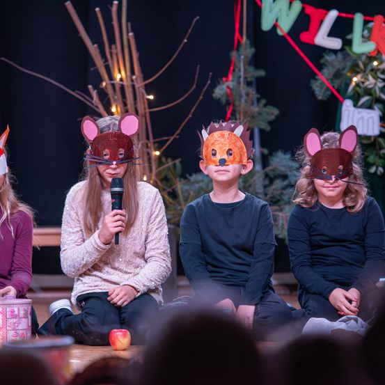 Drei Kinder sitzen auf dem Boden und tragen Tiermasken. Eines hält ein Mikrofon und spricht, während die anderen aufmerksam zuhören. Sie sind von Weihnachtsdekorationen und einem Hintergrund mit einem Banner umgeben.