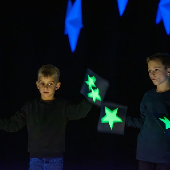 Zwei Jungen in schwarzen Shirts halten beleuchtete Sternausschnitte unter einem dunklen Himmel mit leuchtenden blauen Formen.