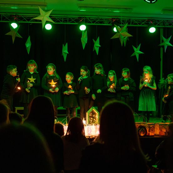 Eine Gruppe von Kindern mit brennenden Kerzen steht auf der Bühne vor einem Publikum, beleuchtet von grünen Lichtern.