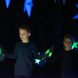 Zwei Kinder in dunklen Shirts halten leuchtende sternförmige Objekte in einem dunklen Raum mit blauen Sternen im Hintergrund.