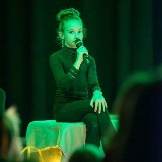 Ein junges Mädchen in einem grünen Langarmshirt und schwarzen Hosen sitzt auf einem Hocker und hält ein Mikrofon in der Hand, wahrscheinlich auf der Bühne unter grünem Licht auftretend.