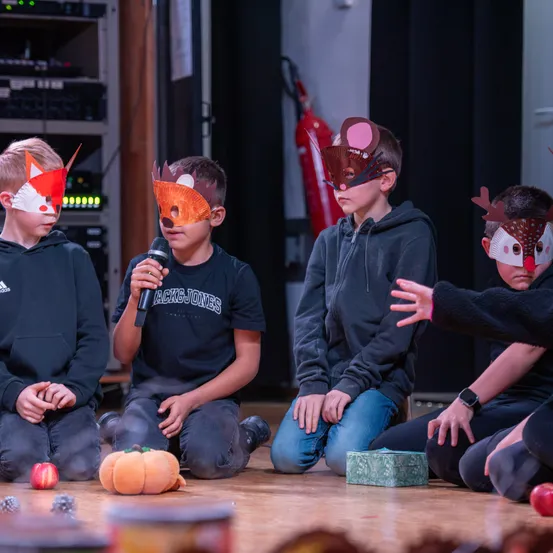 Eine Gruppe von Kindern sitzt auf einer Bühne und trägt Tiermasken. Ein Kind hält ein Mikrofon, während andere zusehen. Eine Kürbis und ein Feuerlöscher liegen auf dem Boden.