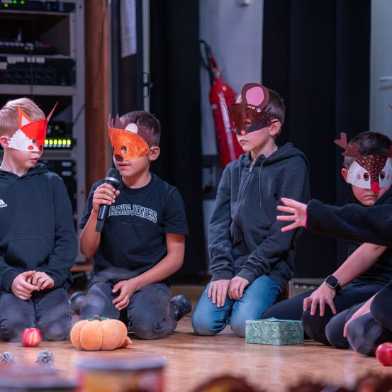 Eine Gruppe von Kindern sitzt auf einer Bühne und trägt Tiermasken. Ein Kind hält ein Mikrofon, während andere zusehen. Eine Kürbis und ein Feuerlöscher liegen auf dem Boden.