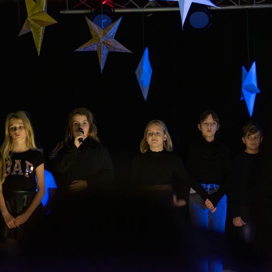 Fünf Kinder stehen auf der Bühne, eines hält ein Mikrofon. Sie sind in Schwarz gekleidet, und blaue Papiersterne hängen über ihnen.