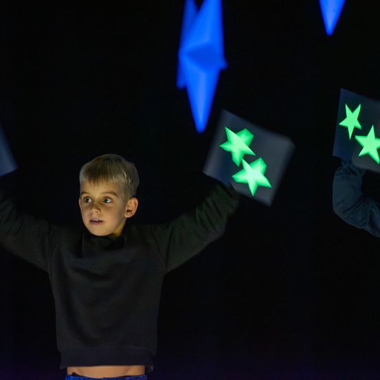 Ein junger Junge in einem schwarzen Shirt hält leuchtende sternförmige Objekte hoch. Er steht in einem dunklen Raum mit blauen Sternen im Hintergrund.
