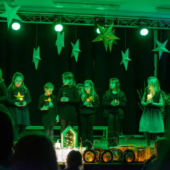 Eine Gruppe von Kindern steht auf der Bühne und hält brennende Kerzen. Sie tragen dunkle Kleidung, und die Bühne ist mit grünen Lichtern und hängenden Sternendekorationen geschmückt.