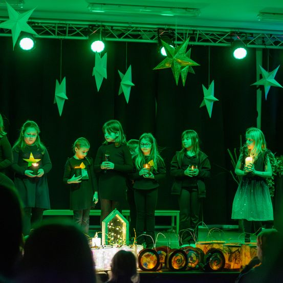 Eine Gruppe von Kindern steht auf der Bühne und hält brennende Kerzen. Sie tragen dunkle Kleidung, und die Bühne ist mit grünen Lichtern und hängenden Sternendekorationen geschmückt.