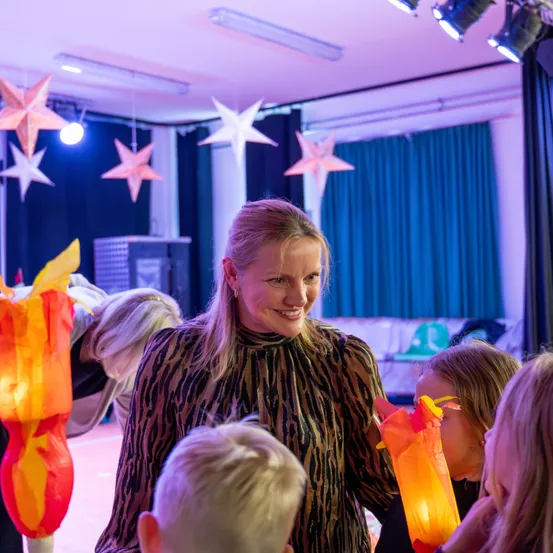 Eine Frau in einem gemusterten Oberteil steht in einem Raum mit Kindern. Sterndekorationen hängen von der Decke. Ein Kind hält eine leuchtende Laterne.