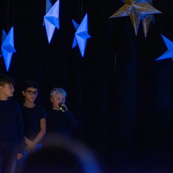 Drei Jungen stehen auf einer Bühne, über ihnen hängen Papiersterne. Ein Junge hält ein Mikrofon.