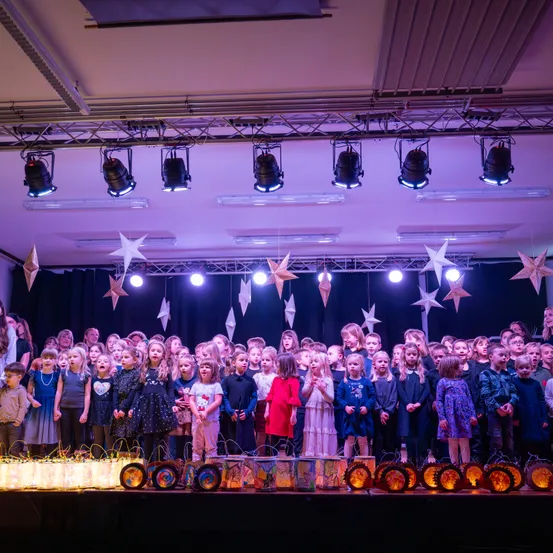 Eine Gruppe von Kindern in Kostümen singt auf einer Bühne mit Sternendekorationen und Lichtern darüber. Sie halten Laternen und stehen vor einer Kulisse mit einem schwarzen Vorhang.