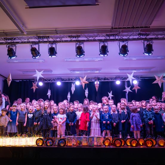Eine Gruppe von Kindern in Kostümen singt auf einer Bühne mit Sternendekorationen und Lichtern darüber. Sie halten Laternen und stehen vor einer Kulisse mit einem schwarzen Vorhang.